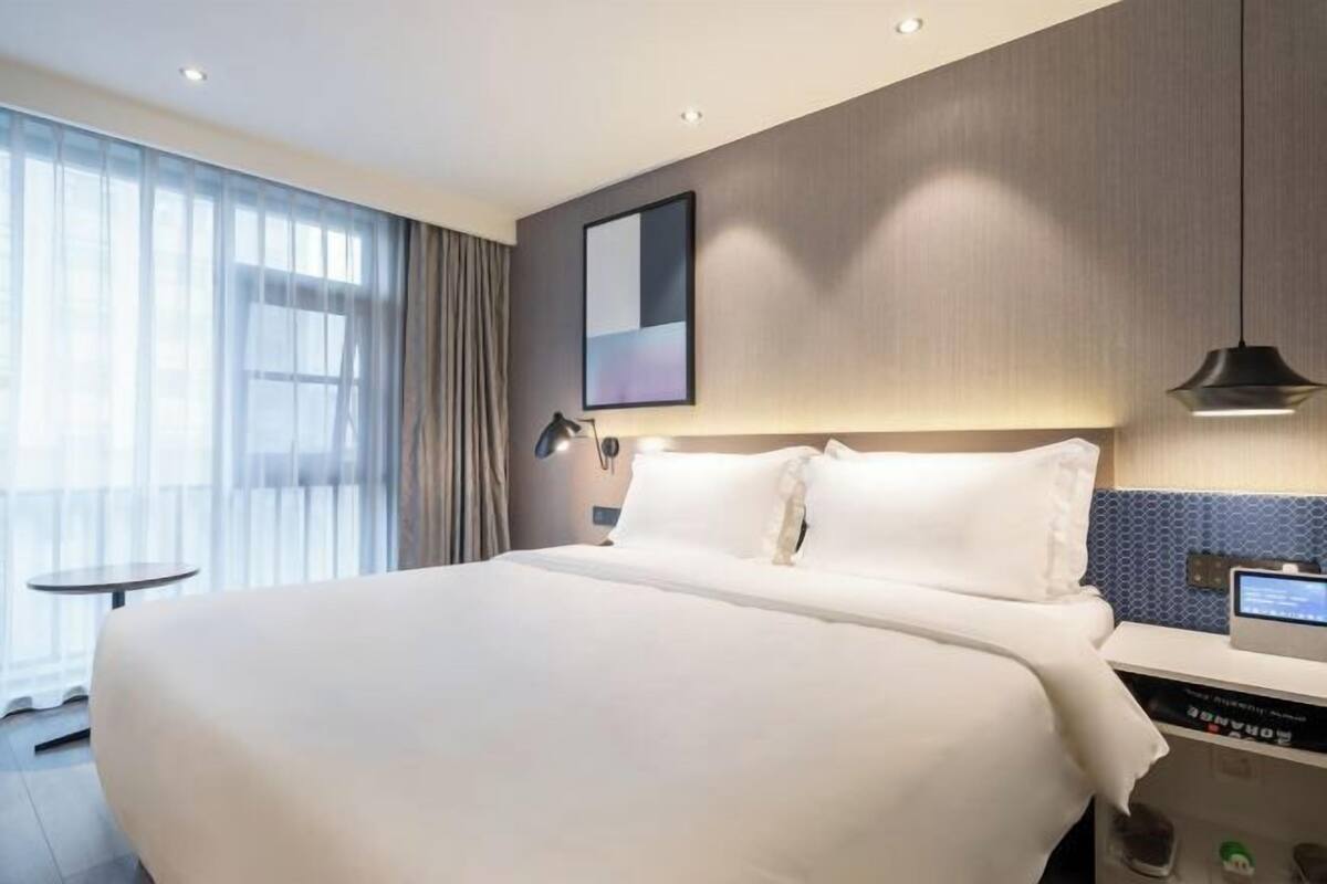 A modern hotel room with a large bed, a bedside table with a digital device, and a view of the outdoors through sheer curtains.