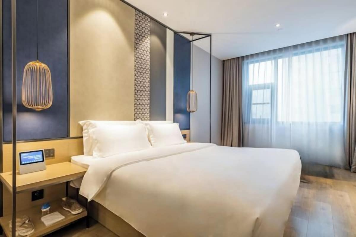 A modern hotel room with a large bed, a bedside table with a control panel, and a large window with curtains.