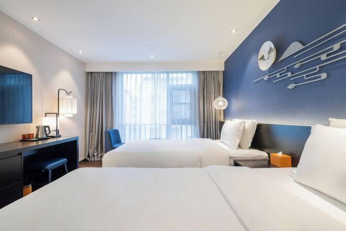 A hotel room with two beds, a desk, a chair, a lamp, and a large window with curtains.
