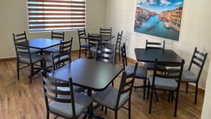 Un comedor con varias mesas y sillas, un ventanal con persianas y un cuadro de paisaje urbano en la pared.