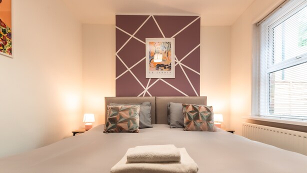 A neatly made bed with pillows, a wall art piece, and a window with blinds. - Monthly Savings - 4 Beds - Sleeps 8 - Free Parking (Southampton)