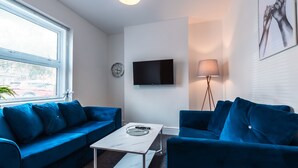 A modern living room with two blue sofas, a white coffee table, a flat-screen TV mounted on the wall, a floor lamp, and a clock on the wall. - Monthly Savings - 4 Beds - Sleeps 8 - Free Parking (Southampton)