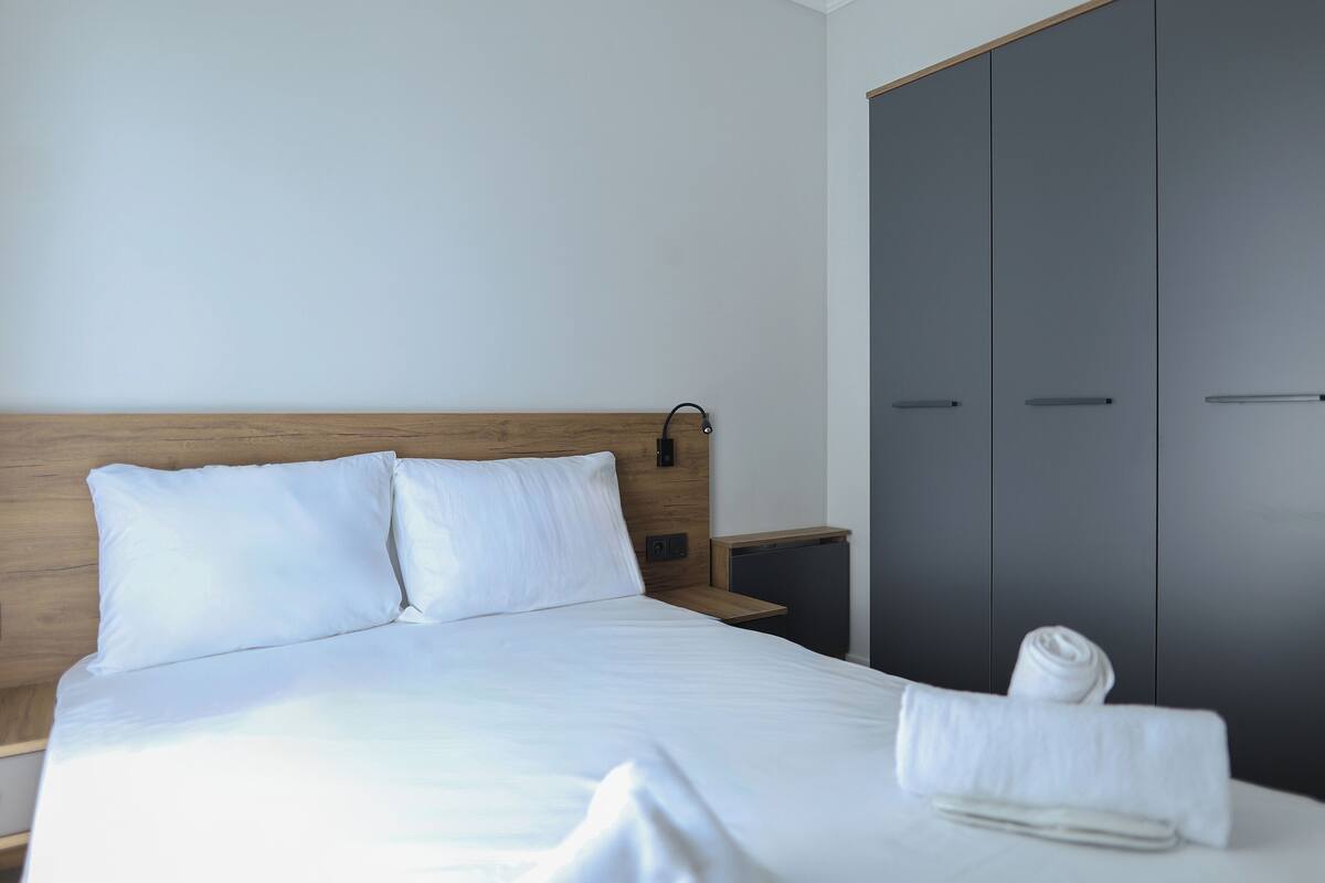 A neatly made bed with white linens, a wooden headboard, a nightstand with a lamp, and a grey wardrobe.