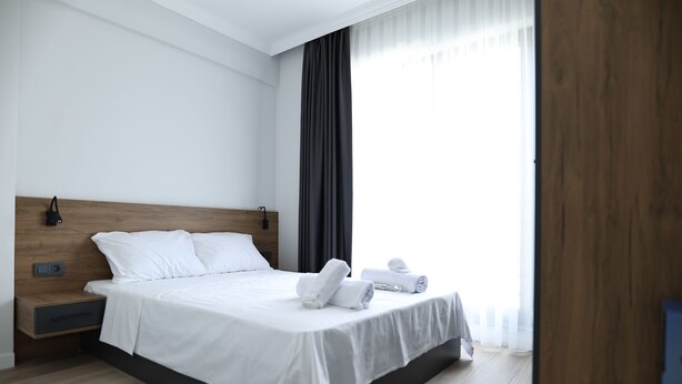 A modern hotel room with a wooden headboard, white bedding, a bedside table with a lamp, and a large window with sheer curtains. - Deluna Yaşamkent (Ankara)
