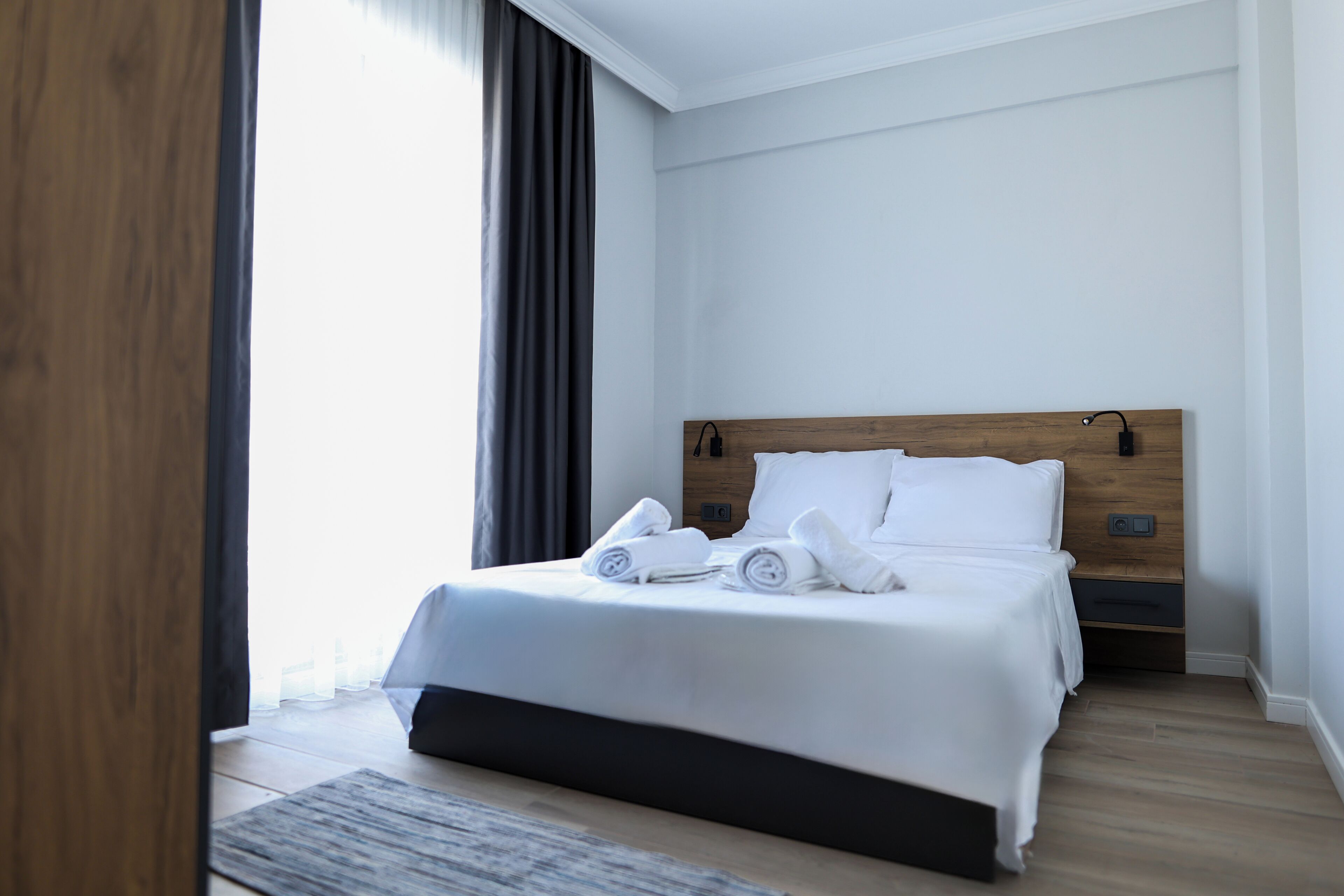 A modern hotel room with a wooden headboard, a neatly made bed with white linens and towels, and a large window with curtains.