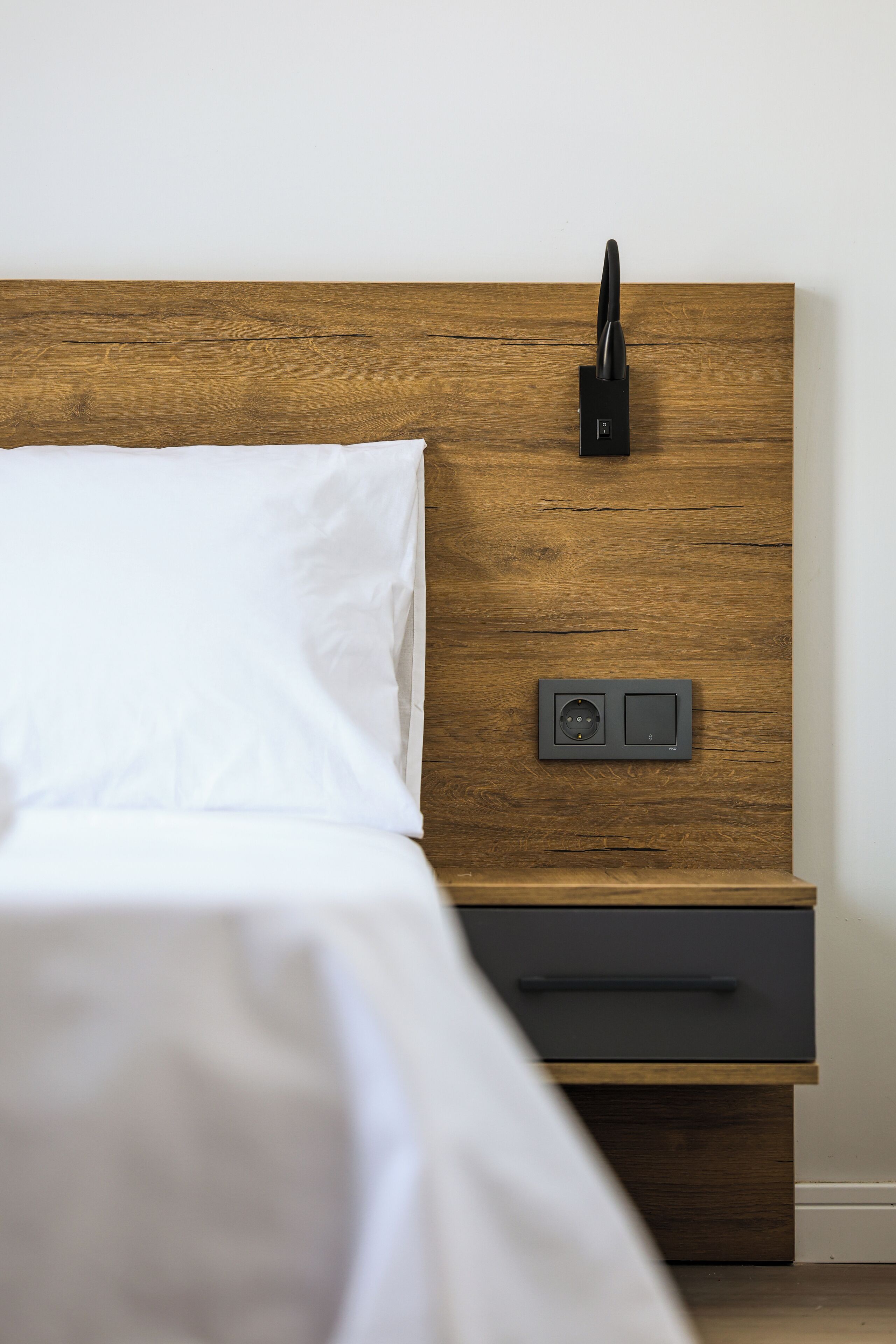 A neatly made bed with white linens, a wooden headboard, a bedside table with a drawer, and a small electronic device on the headboard.