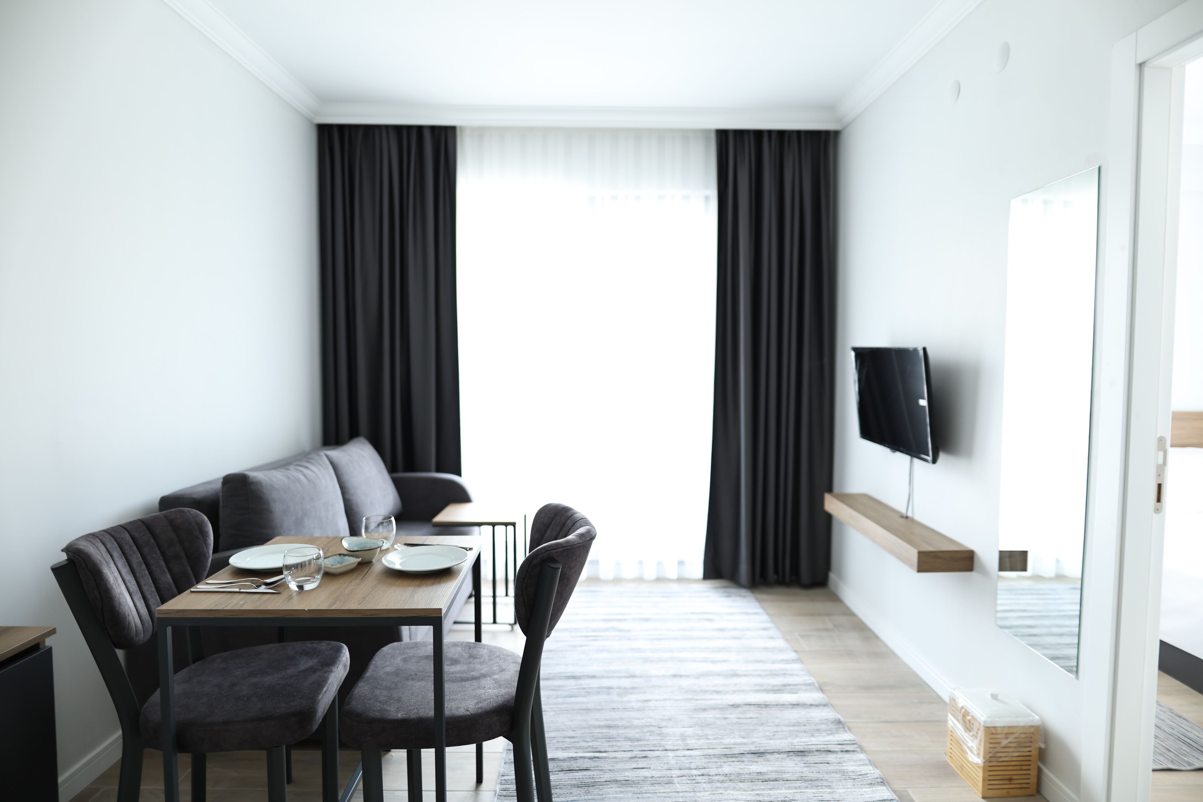 Een moderne, minimalistische woonkamer met een eethoek, met een bank, twee stoelen, een houten tafel en een flatscreen televisie aan de muur bevestigd.