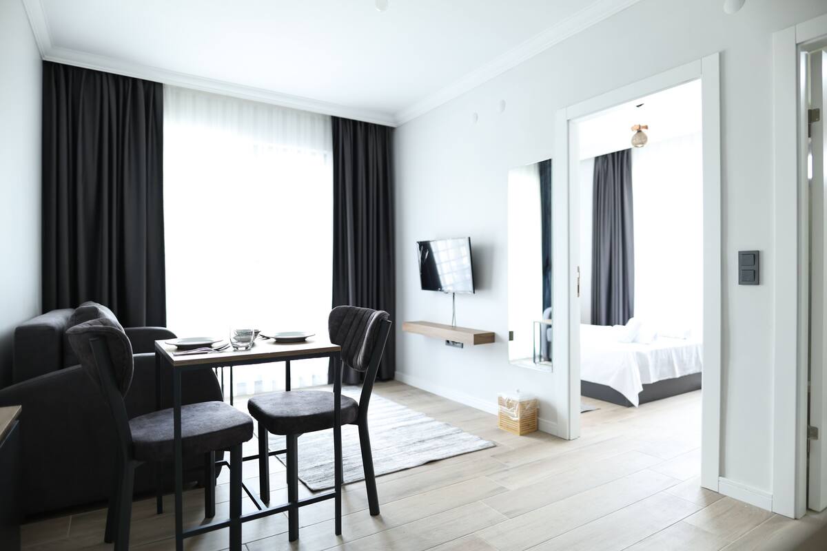 A modern hotel room with a dining area, a TV mounted on the wall, and a bed with a view of the outdoors.