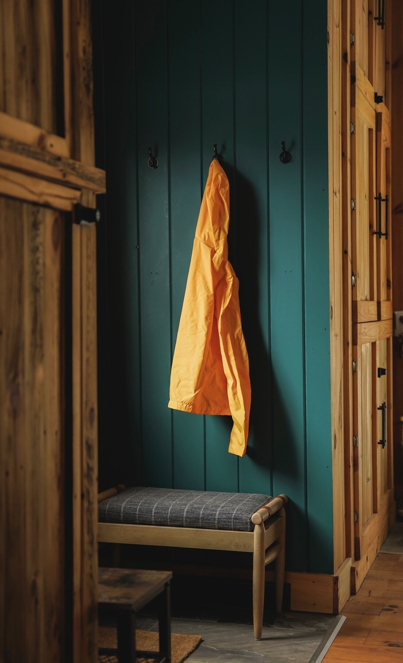 A wooden bench with a cushion against a teal wall, a wooden door with a handle, and a yellow jacket hanging on the wall.
