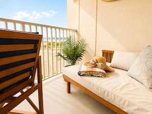 A balcony with a wooden chair, a bed with a striped blanket, a potted plant, and a view of the beach. - Oceanfront Condo | NEW Retro Design | Beach + Pool (Wildwood)