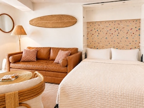 Une chambre avec un canapé en cuir marron, une table basse ronde en osier, un lit blanc avec une tête de lit à motifs et un miroir fixé au mur.