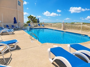 Une piscine à l’eau bleue, entourée de chaises longues blanches et de parasols, avec un ciel dégagé et des arbres qui se dessinent à l’horizon.