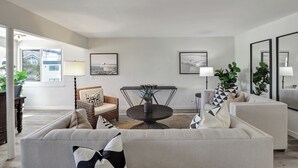 Un salon moderne avec un canapé, des fauteuils, une table basse ronde et une table à manger. Les murs sont ornés de tableaux encadrés et il y a des plantes en pot.