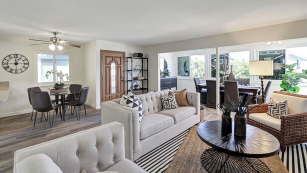 Un salon spacieux comprenant un canapé, un fauteuil, une table à manger et des chaises. On y trouve également un ventilateur de plafond, une horloge et une fenêtre donnant sur l’extérieur.
