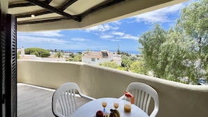 Un balcon avec une table dressée pour deux personnes, une bouteille de jus de fruits et une assiette de fruits. Le balcon offre une vue sur une ville côtière, avec ses bâtiments et ses arbres.