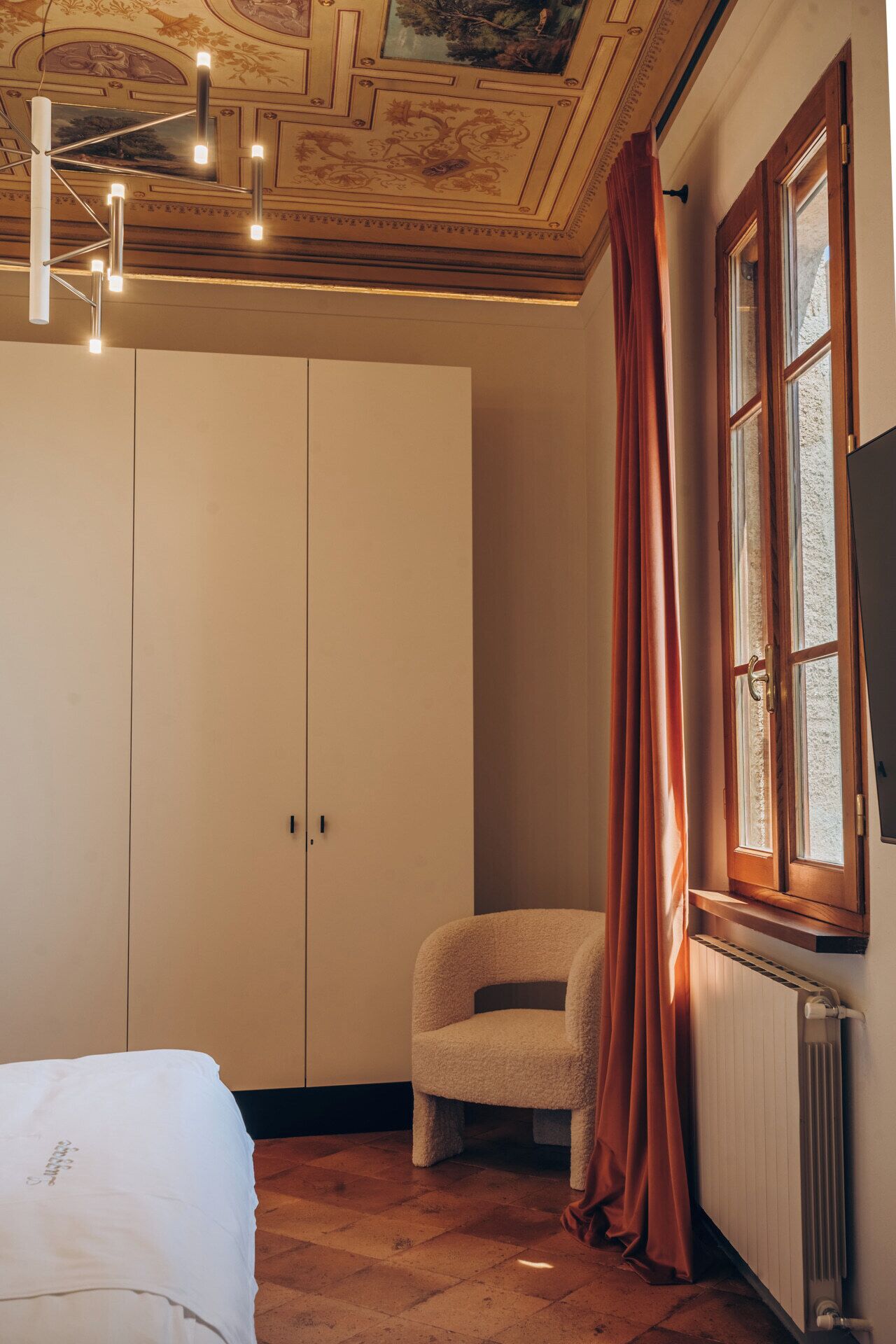 A bedroom with a large wardrobe, a chair, a radiator, and a window with curtains.