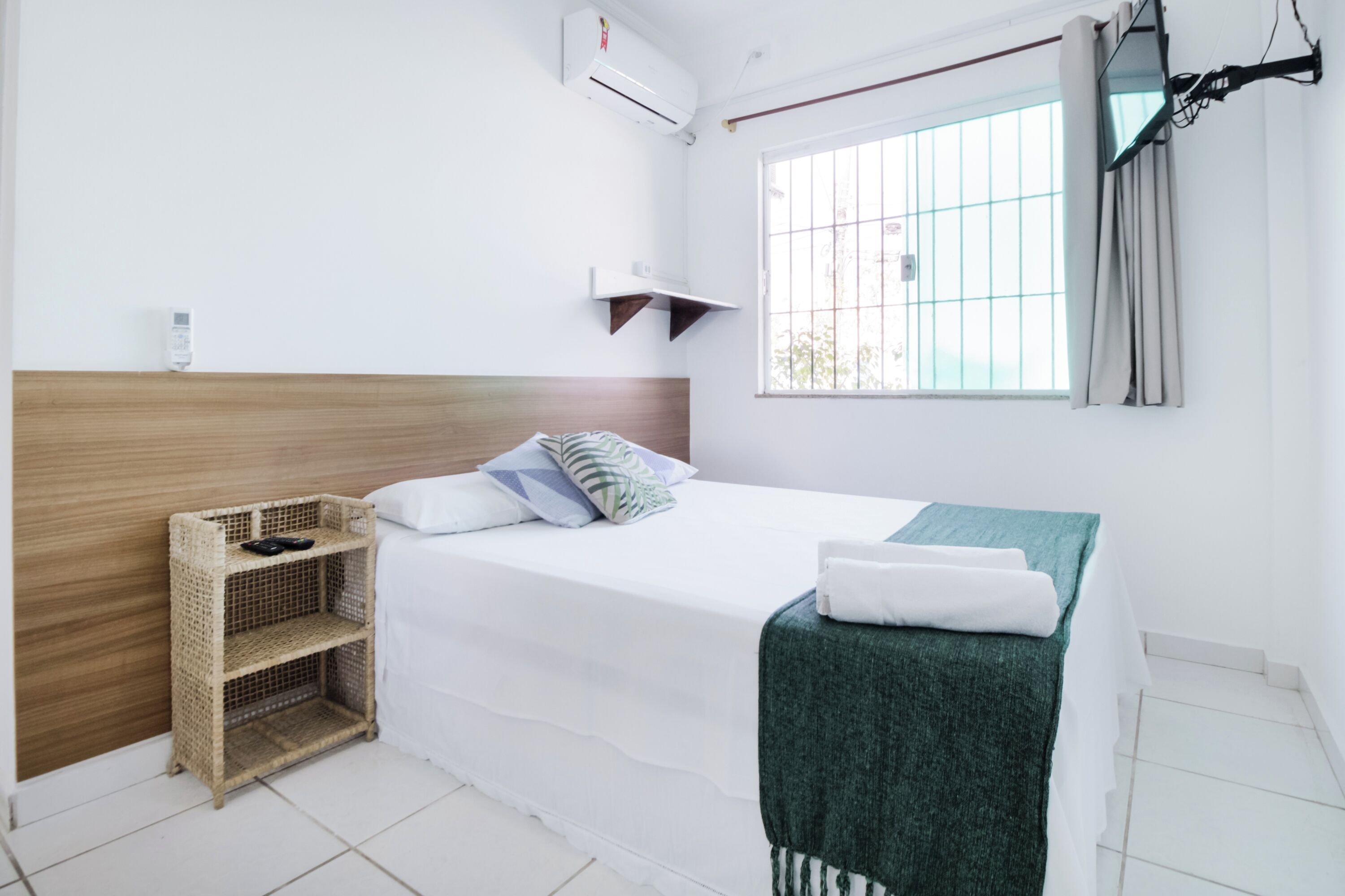 A clean, modern hotel room with a wooden headboard, a single bed with a green blanket, a wicker side table, and a window with a view of the outside.