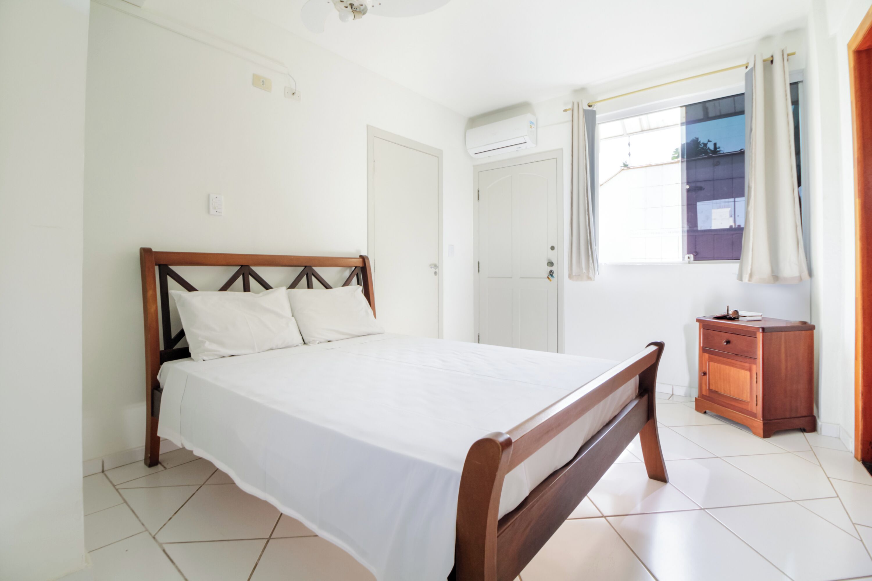 A bedroom with a wooden bed, a small wooden nightstand, a window with curtains, and an air conditioning unit on the wall.