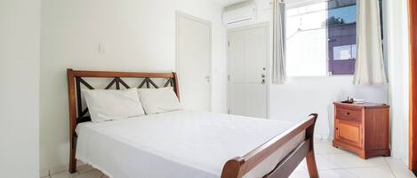 A bedroom with a wooden bed, a small wooden nightstand, a window with curtains, and an air conditioning unit on the wall.
