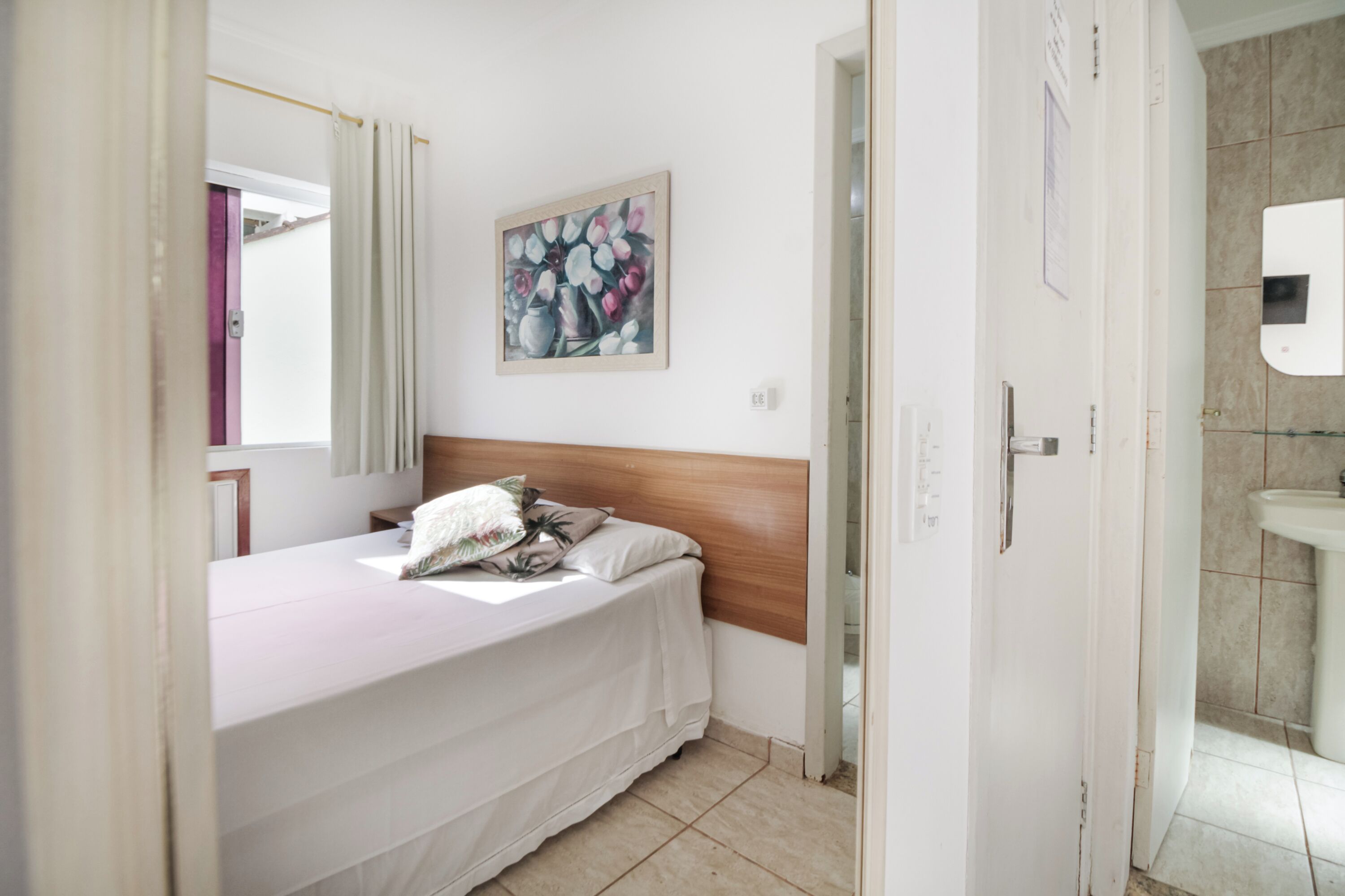 Un dormitorio con cama, cabecera de madera, un cuadro en la pared, baño con lavamanos y espejo, y una puerta que da a otra habitación.