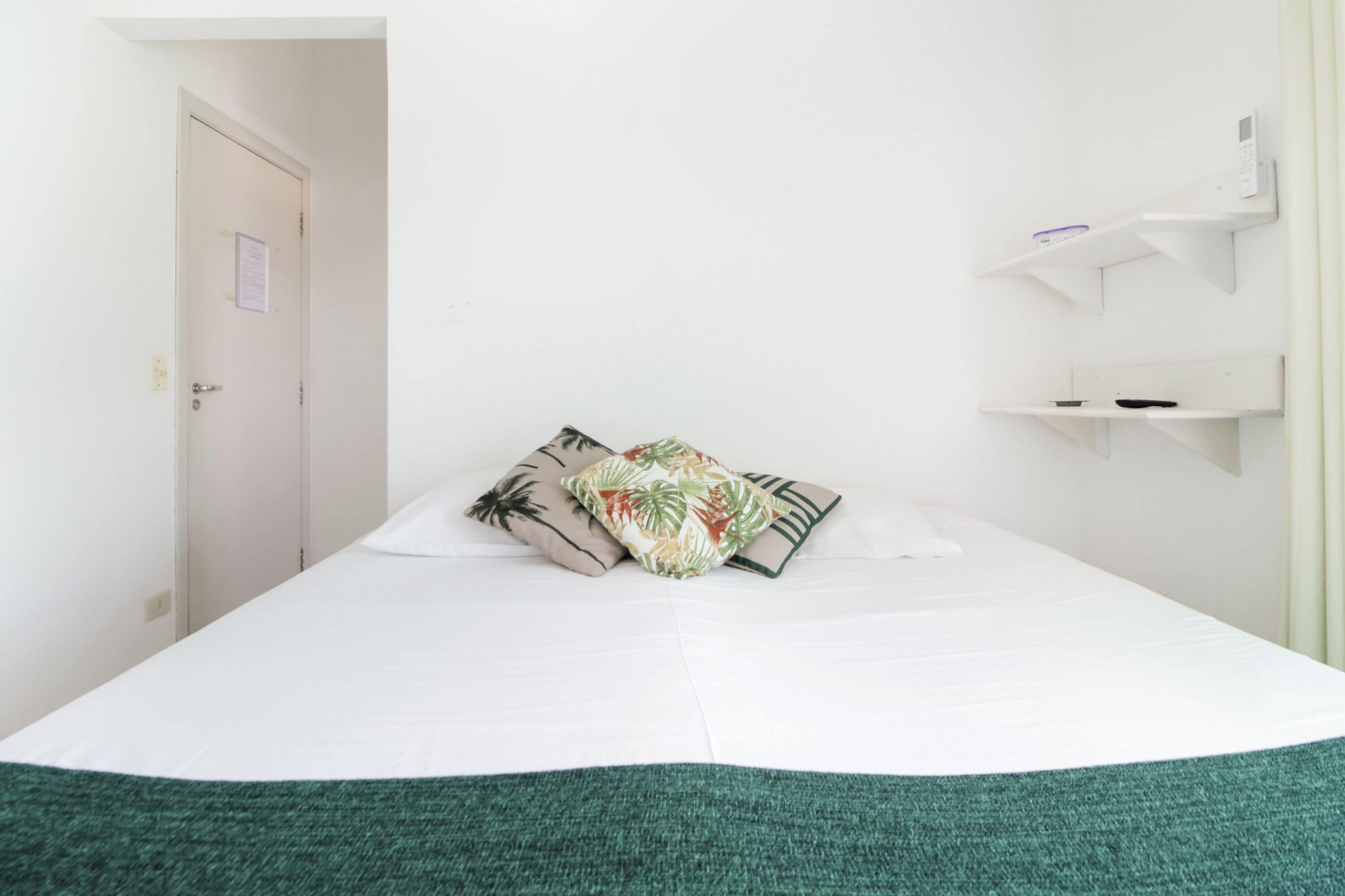 A hotel room with a bed, a door, and a wall-mounted shelf with a small item on it.