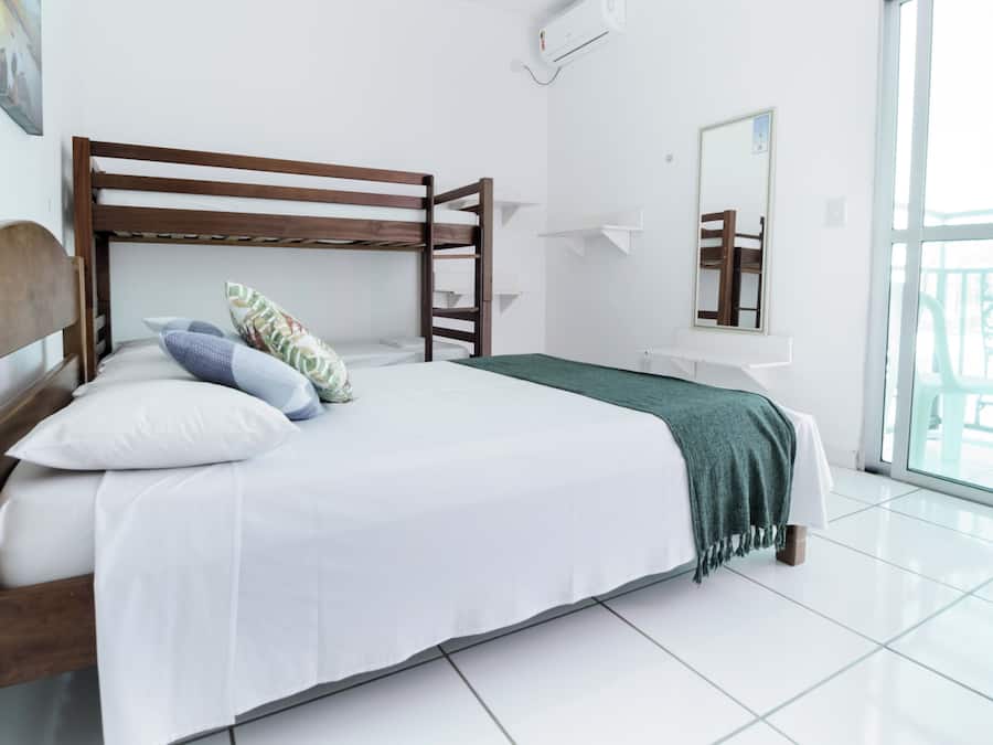 Un dormitorio con una cama litera de madera, ropa de cama blanca, un edredón verde, un ventanal con vistas y un equipo de aire acondicionado blanco.