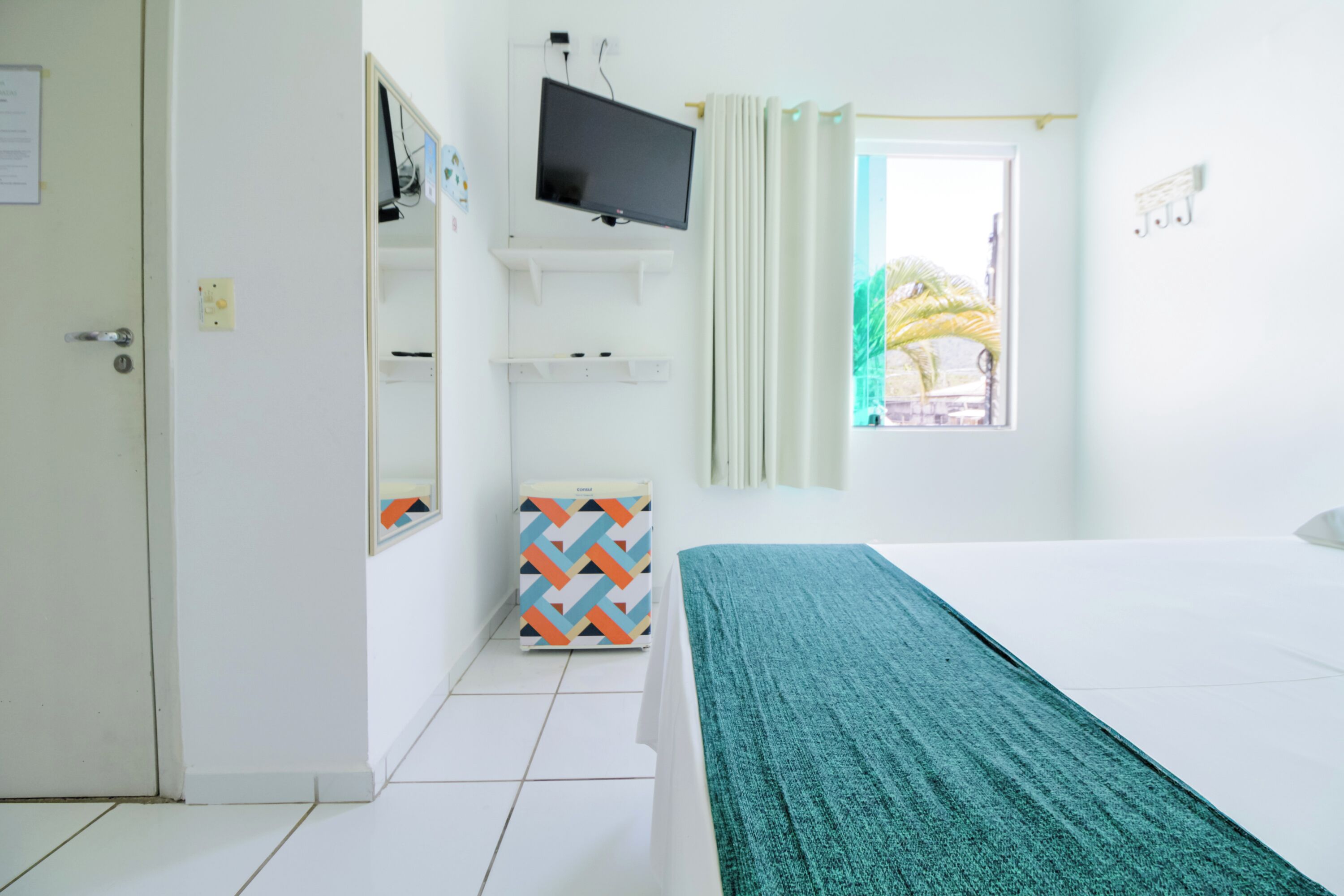 A hotel room with a bed, a television mounted on the wall, a window with curtains, and a small cabinet with a geometric-patterned box.