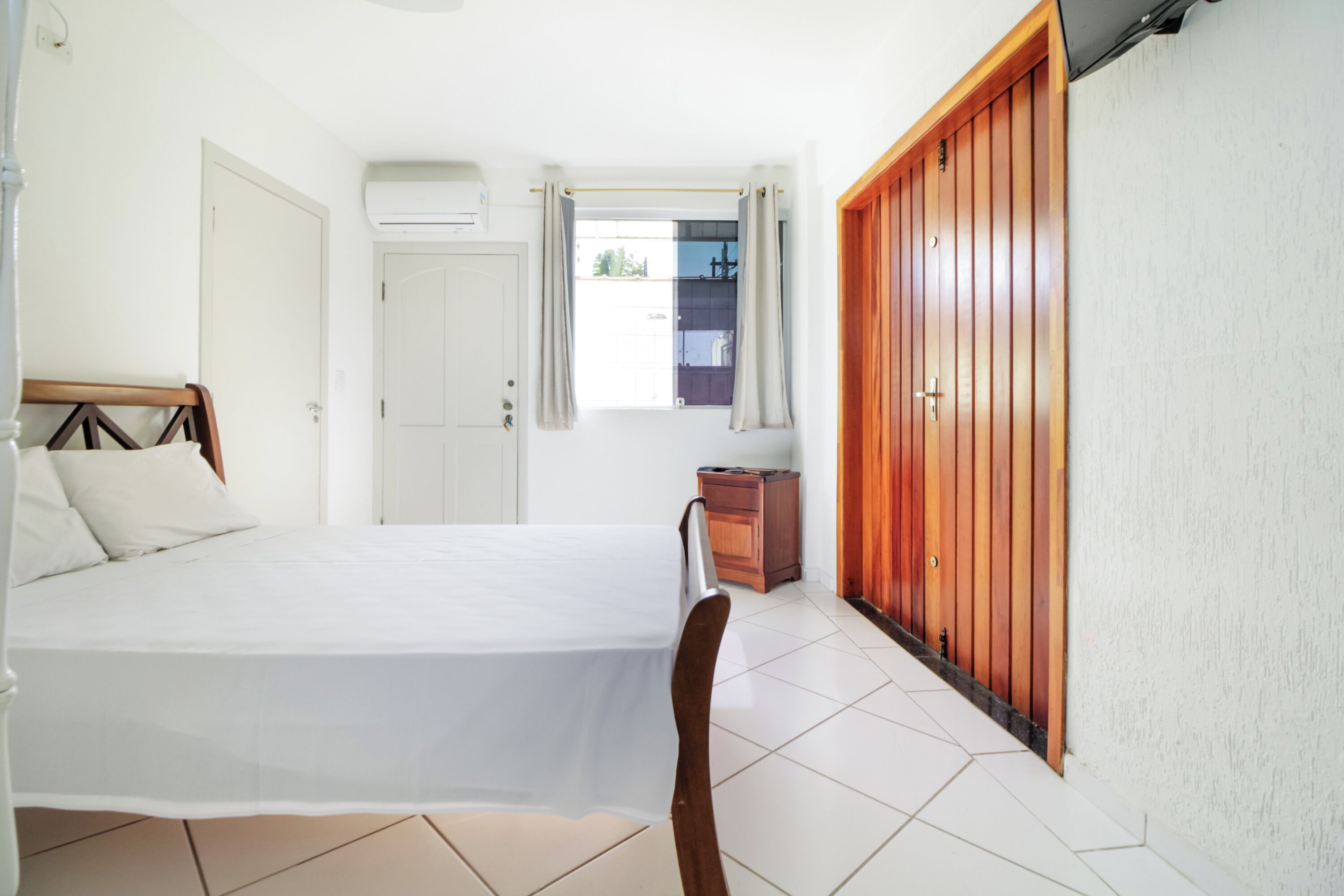 Un dormitorio con cama, armario de madera, ventana con cortinas y una mesita de noche pequeña.