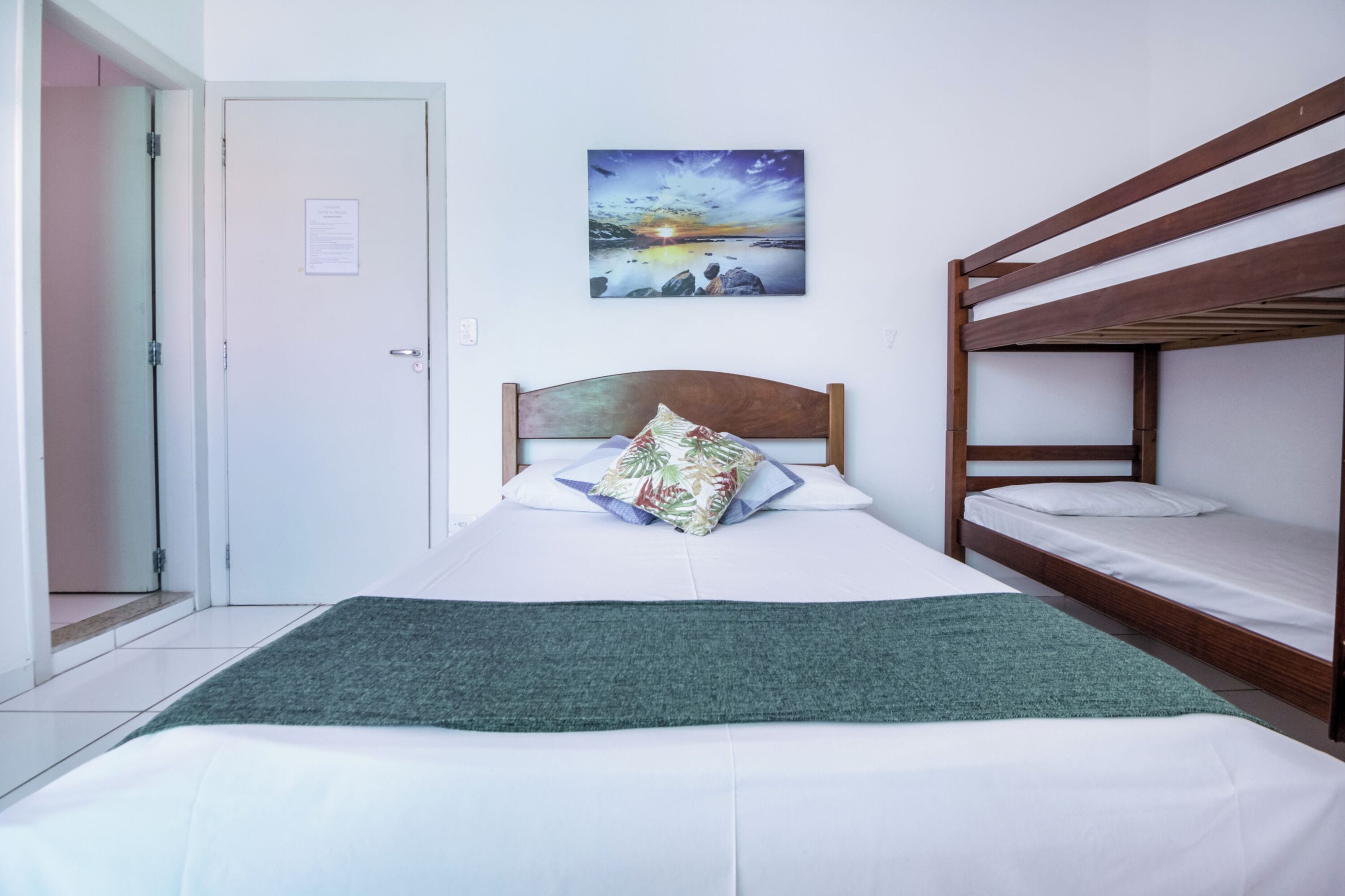 Een enkel bed met houten hoofdbord, een groene dekensprei en een bloemenkussen. Een stapelbed staat rechts. Een schilderij van een zonsondergang boven water hangt aan de muur.