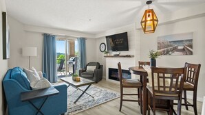 A modern living room with a blue sofa, a dining table with wooden chairs, a fireplace, a TV mounted on the wall, and a view of the outdoors through large windows.
