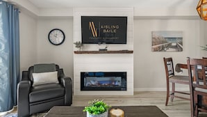A living room with a fireplace, a television mounted above it, a clock, and a dining area with chairs.