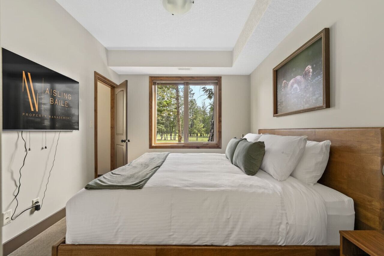 Une chambre avec un grand lit, un téléviseur fixé au mur, une fenêtre donnant sur les arbres et un tableau encadré accroché au mur.