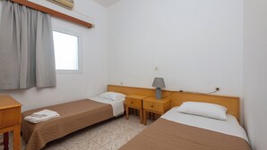 A room with two single beds, a wooden nightstand with a lamp, a window with curtains, and an air conditioning unit. - Seafront apartment in the bay of Kefalos, Kos (Kefalos)