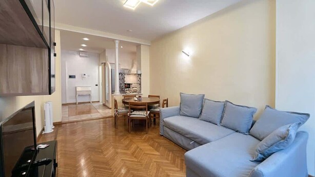 A modern living room with a grey sofa, a dining table with chairs, and a kitchen area in the background. - Center Apartment - Affitti Brevi Italia (Lecco)