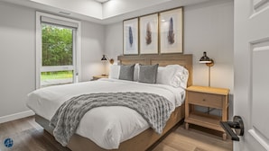 Ein modernes Schlafzimmer mit einem großen Bett, einem Nachttisch, einem Fenster mit Blick auf Grünflächen und Wandbildern.