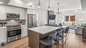A modern kitchen with stainless steel appliances, a central island with bar stools, and a dining area with a gray sofa and a TV mounted on the wall.