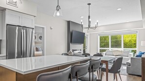 A modern kitchen with a central island, bar stools, a dining area with a table and chairs, a flat-screen TV mounted on the wall, and a large window offering a view of greenery. - 2 Bed/2 Bath Sparrowhawk Resort-Sleeps 6 (Dead Man's Flats)