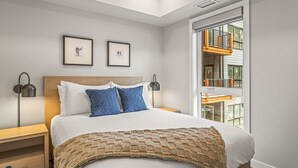 Ein modernes Schlafzimmer mit einem großen Bett, zwei gerahmten Bildern an der Wand, einem Nachttisch mit Lampe und einem Fenster mit Blick auf ein Gebäude.