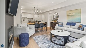 A modern living room with a sofa, ottoman, and coffee table, a dining area with chairs, and a kitchen with cabinets and a TV. - The Westview - 2 Bed/2Bath Sleeps 6 (Dead Man's Flats)