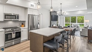 A modern kitchen with white cabinets, stainless steel appliances, a central island with bar stools, and a large window offering a view of greenery.