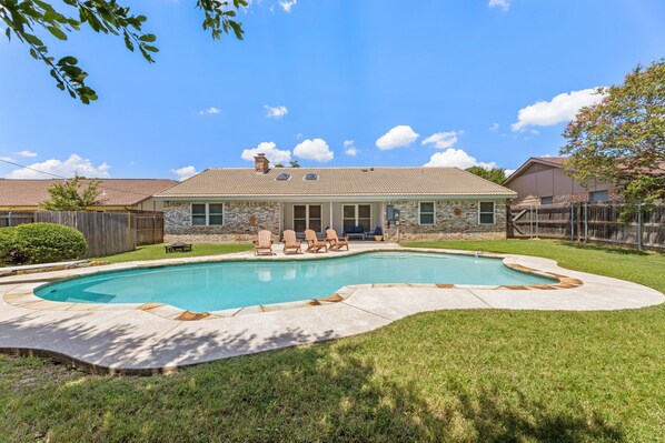 A backyard with a swimming pool, a patio with chairs, and a house with a tiled roof.
