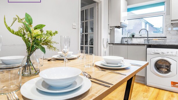 Een moderne keuken met een ingebouwde wasmachine en droger, een eettafel gedekt met witte borden en glazen, en een vaas met groene planten.