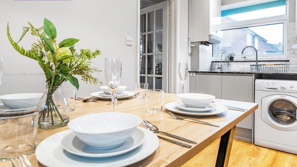 Uma cozinha moderna com máquina de lavar e secadora embutidas, uma mesa de jantar posta com pratos e copos brancos, e um vaso com plantas verdes.