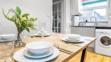 A modern kitchen with a built-in washer and dryer, a dining table set with white plates and glasses, and a vase with green plants.