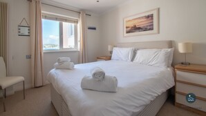 A hotel room with a large bed, a bedside table with a lamp, a chair, and a window with curtains. - Pebbles - The Haven, Swanage (Swanage)