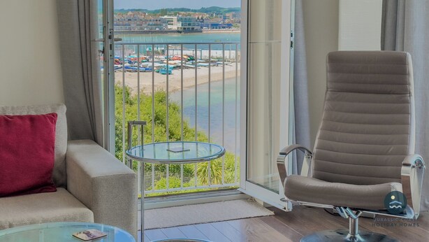 A modern living room with a view of a beach and marina, a glass table, a cushioned sofa, and a reclining chair. - Pebbles - The Haven, Swanage (Swanage)