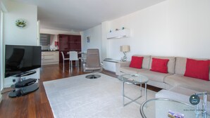 A modern living room with a flat-screen TV, a glass coffee table, a beige sofa with red cushions, and a dining area in the background. - Pebbles - The Haven, Swanage (Swanage)