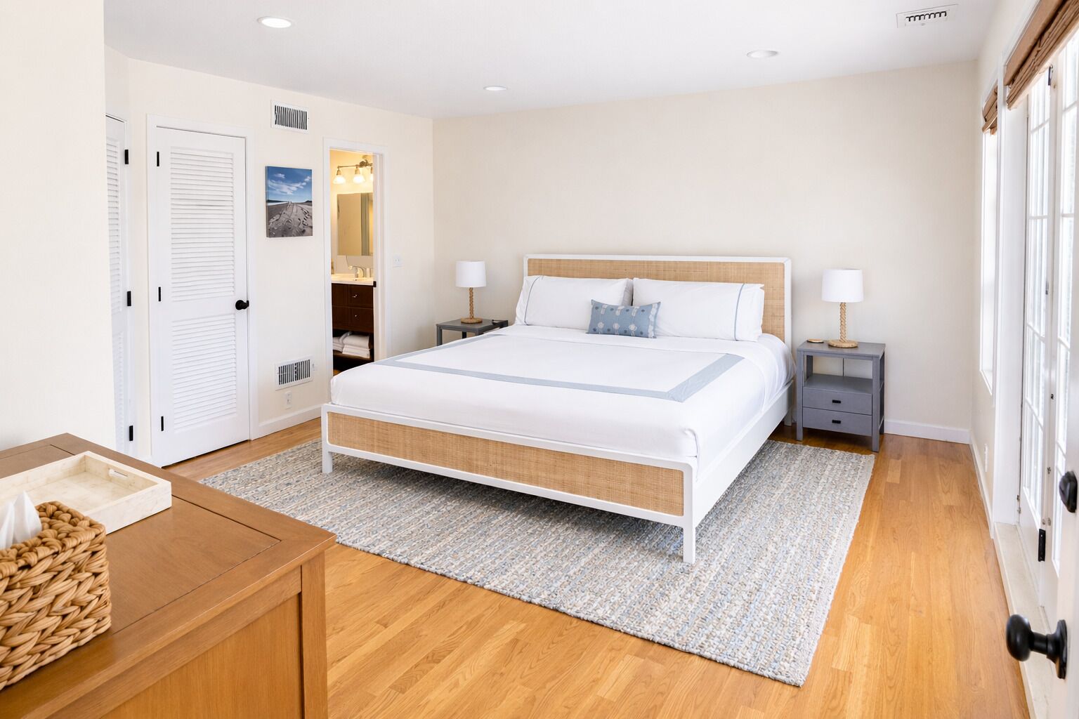 A bedroom with a bed, bedside tables, a nightstand, a wooden dresser, a woven basket, and a door.