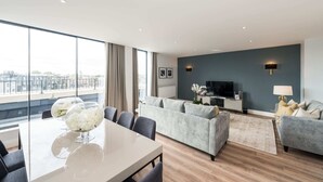 A modern living room with a grey sofa, a dining table with chairs, and a TV on a stand.