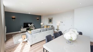 A modern living room with a grey sofa, a dining table with chairs, and a TV on a stand.